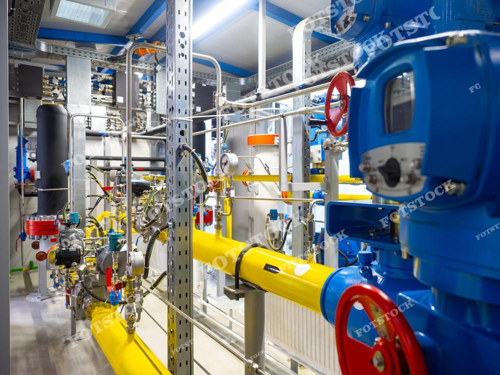Фотография инженерной газовой установки, состоящей из желтых труб, синих управляющих узлов и красных клапанов. Система оборудована манометрами и регулирующими элементами для точного контроля и управления потоком газа в производственном помещении.